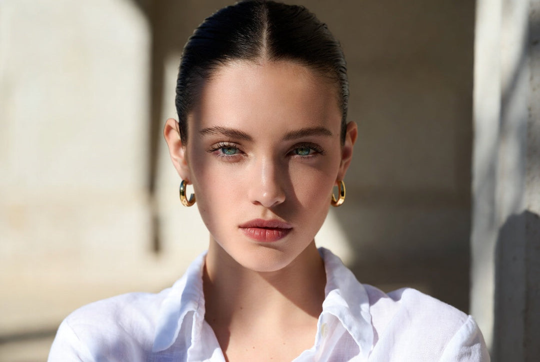 Woman wearing minimalist gold hoop earrings with a white linen shirt in soft sunlight.