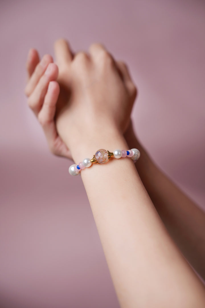 A hand wearing a Elegant handmade beaded bracelet featuring a pink hand-blown glass Liuli bead with gold foil and blue Eastern floral ink splatters, accented with white pearls and pink quartz beads.