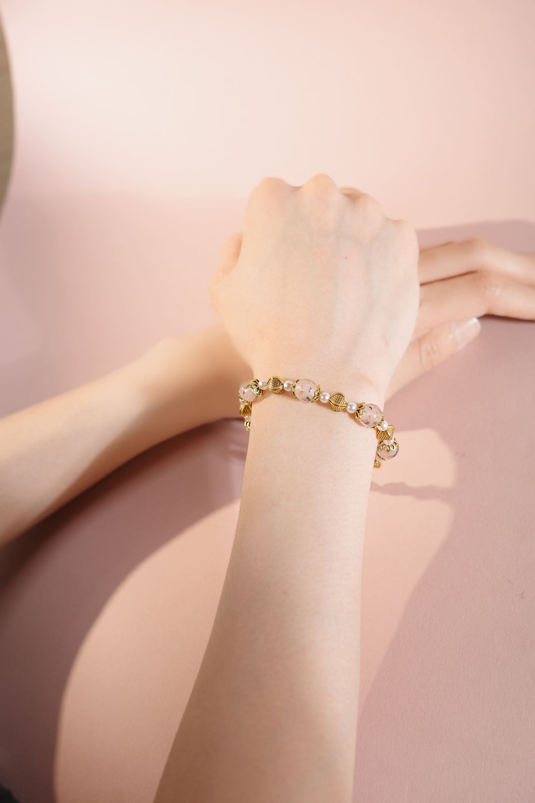 Pink floral Liu Li pearl bracelet with gold accents on wrist, pink background