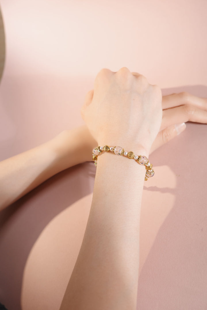 Pink floral Liu Li pearl bracelet with gold accents on wrist, pink background